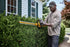 Man trimming bushes beside a house with Dewalt DCHT870B 60V MAX Cordless Hedge Trimmer