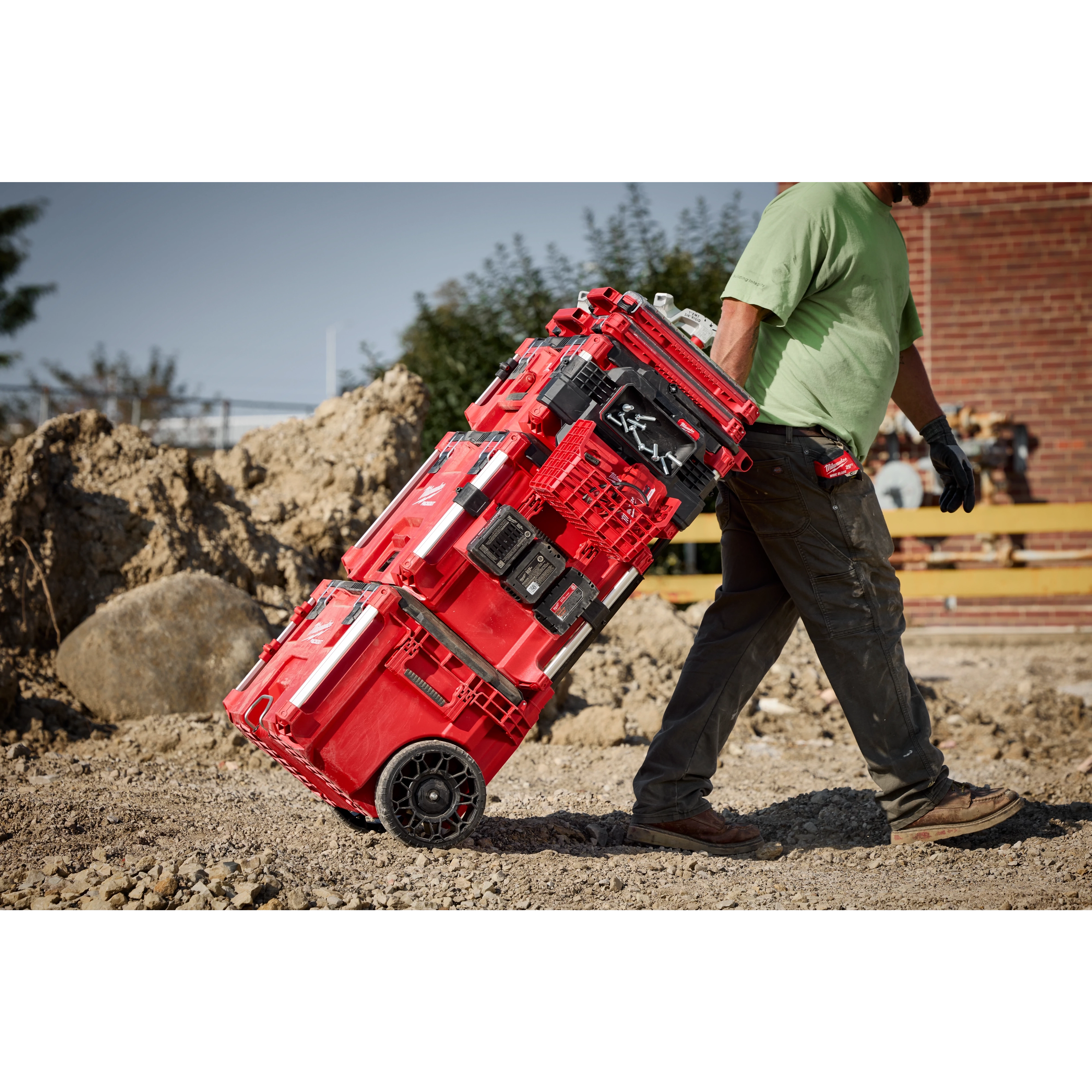 Milwaukee PACKOUT Rolling Tool Box being pulled across a rugged jobsite with stacked PACKOUT components